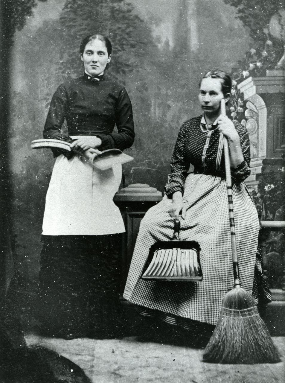 A rectangular image of two women sitting. The one to the left holds a dustpan and the one on the right holds a broom and dustpan.