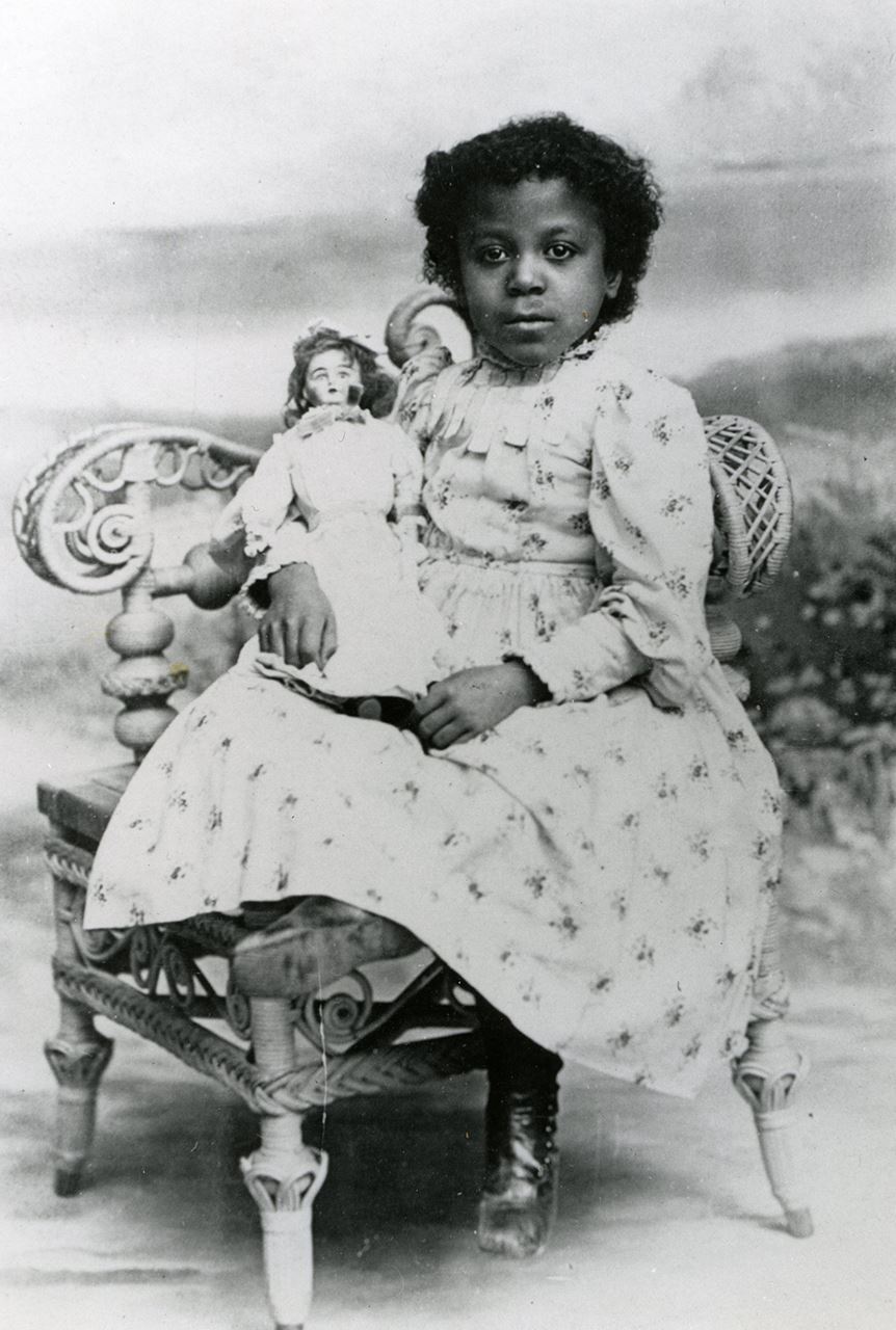 A rectangular black and white image of a young girl sitting in a chair while holding a doll.