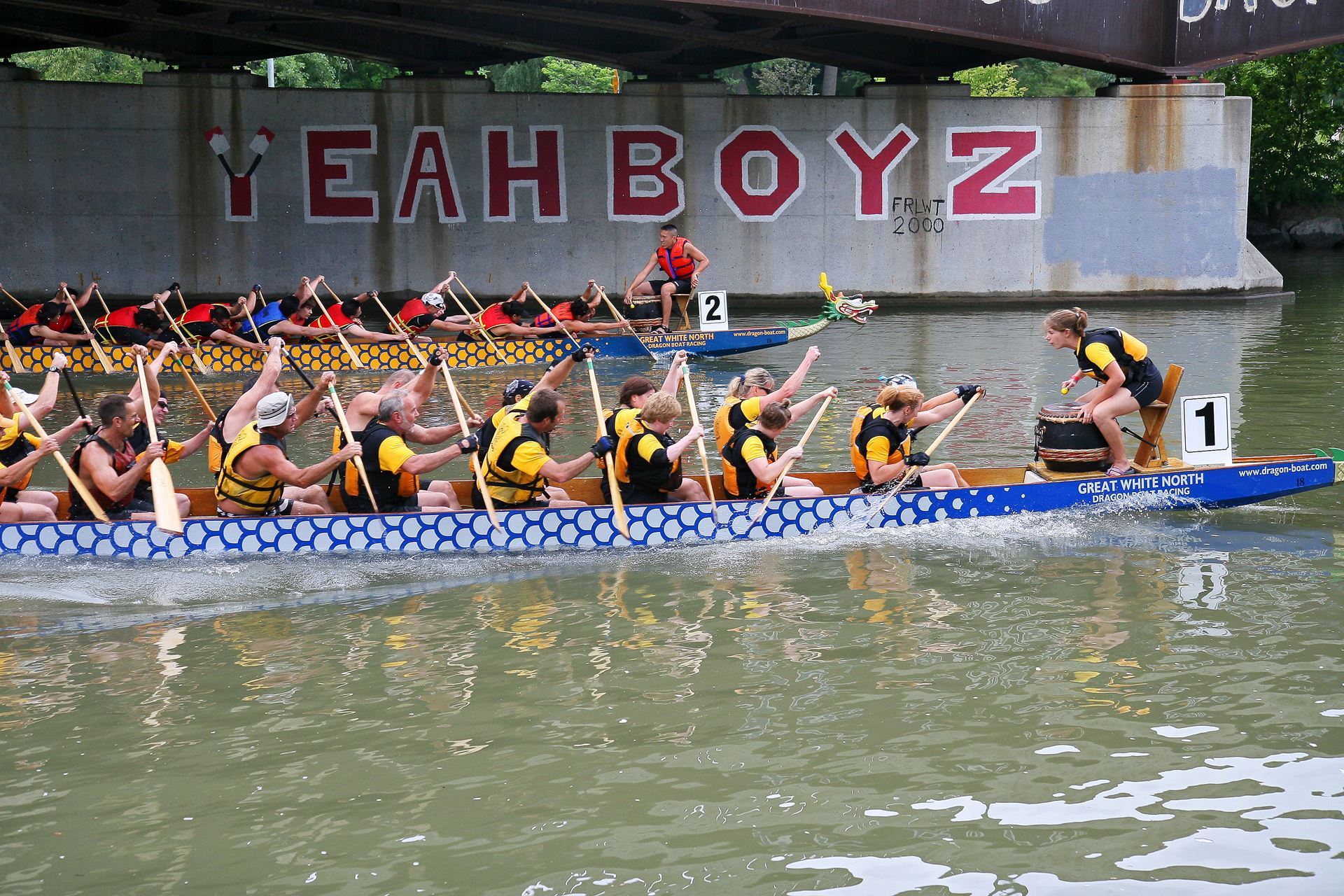 Image of two dragon boats labelled 1 and 2 racing under a bridge 