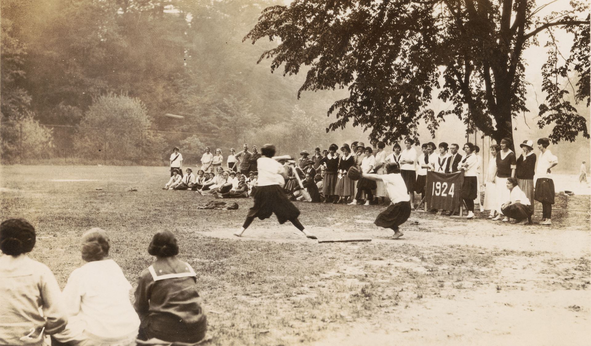 Image of a baseball game from 1924. The batter is about to hit an incoming ball, and a crowd is watching holding a sign reading 1924.