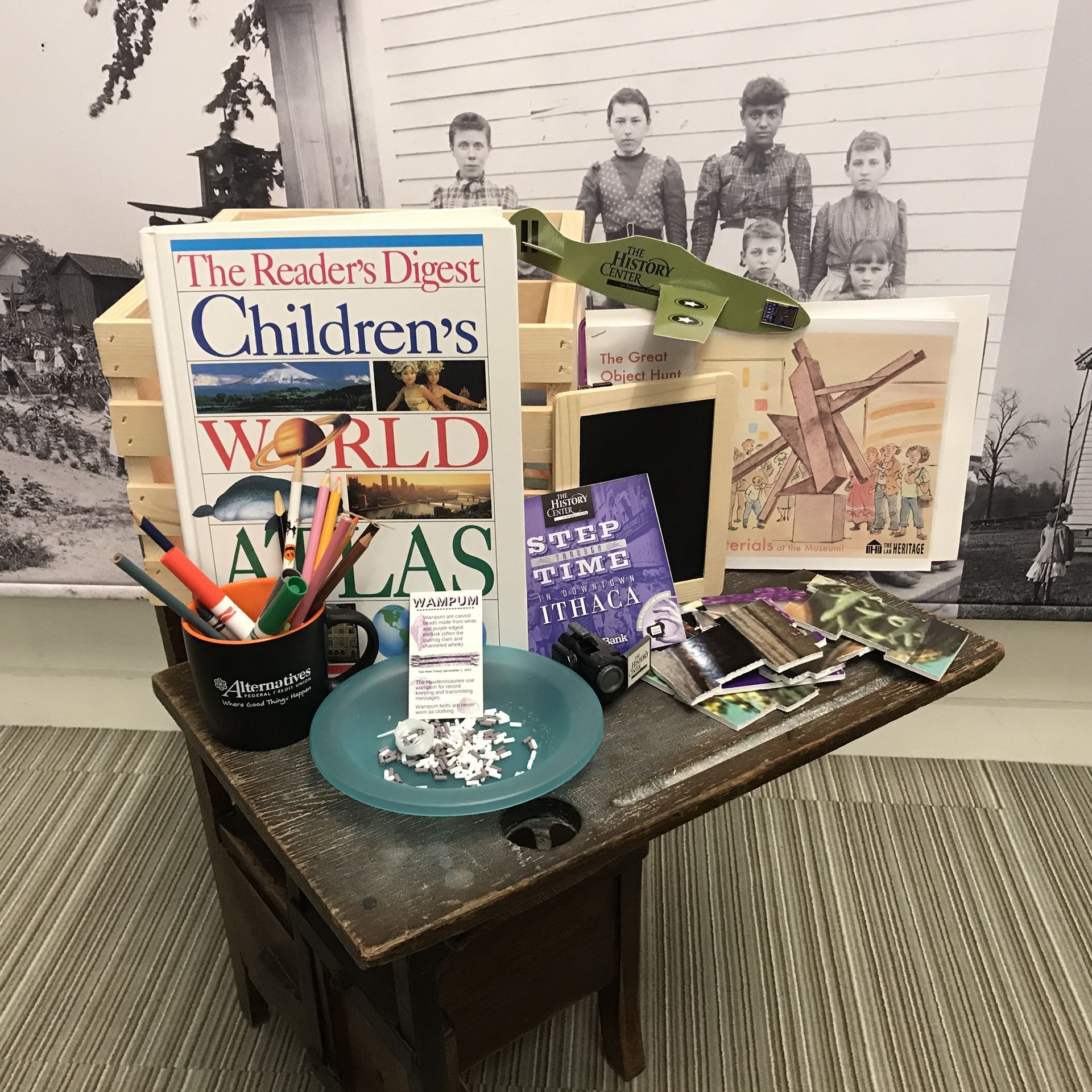 A photo of a museum activity crate, surrounded by a large children's atlas, a cup full of writing utensils. and other various activities.